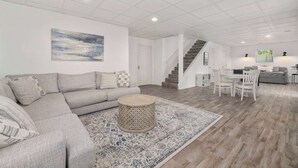 Ein großzügiger Wohnraum mit einem grauen Ecksofa, einem runden Holztisch und einem gemusterten Teppich. Eine Treppe führt auf eine obere Ebene.
