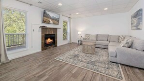Ein geräumiges Wohnzimmer mit Kamin, einer Eckcouch, einem runden Couchtisch und einem großen Fenster mit Jalousien.