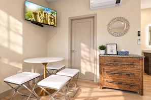 Une pièce avec un téléviseur fixé au mur, un miroir rond, un meuble en bois avec des tiroirs et une table ronde entourée de quatre chaises blanches à coussin.