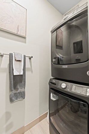 Une buanderie équipée d’un lave-linge et d’un sèche-linge superposés, d’un porte-serviettes avec des serviettes et d’une œuvre d’art encadrée accrochée au mur.