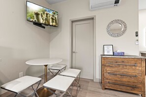 A modern room with a flat-screen TV, a round table with four chairs, a wooden dresser, a door, and a decorative mirror.