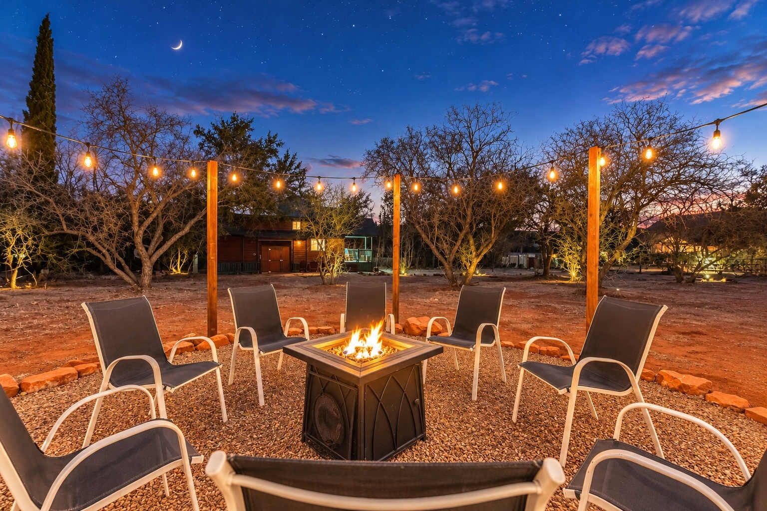 An outdoor seating area with a fire pit, surrounded by chairs and string lights against a night sky.