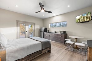 A bedroom with a bed, a ceiling fan, a TV, a kitchen area, and a window.