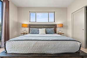 A large bed with a quilted mattress, two bedside tables with lamps, a window above the bed, and a door to the right.
