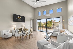 A modern hotel room with a dining area, a sofa, a ceiling fan, and large windows offering a view of the outdoors.