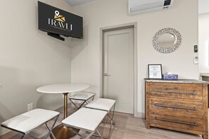 Camera d'albergo con TV a schermo piatto, uno specchio rotondo, una cassettiera in legno e un tavolino con due sedie.