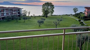 A balcony with a view of a grassy area, a large tree, and a body of water with buildings in the background. - Specchio sul Lago (Manerba del Garda)
