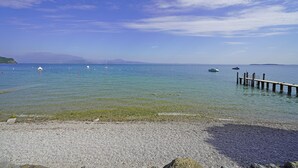 A pebbled beach with a wooden pier extending into calm waters, boats, and distant mountains under a clear sky. - Specchio sul Lago (Manerba del Garda)
