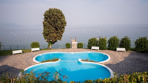A swimming pool with a stone deck, benches, and a view of the sea. - Specchio sul Lago (Manerba del Garda)