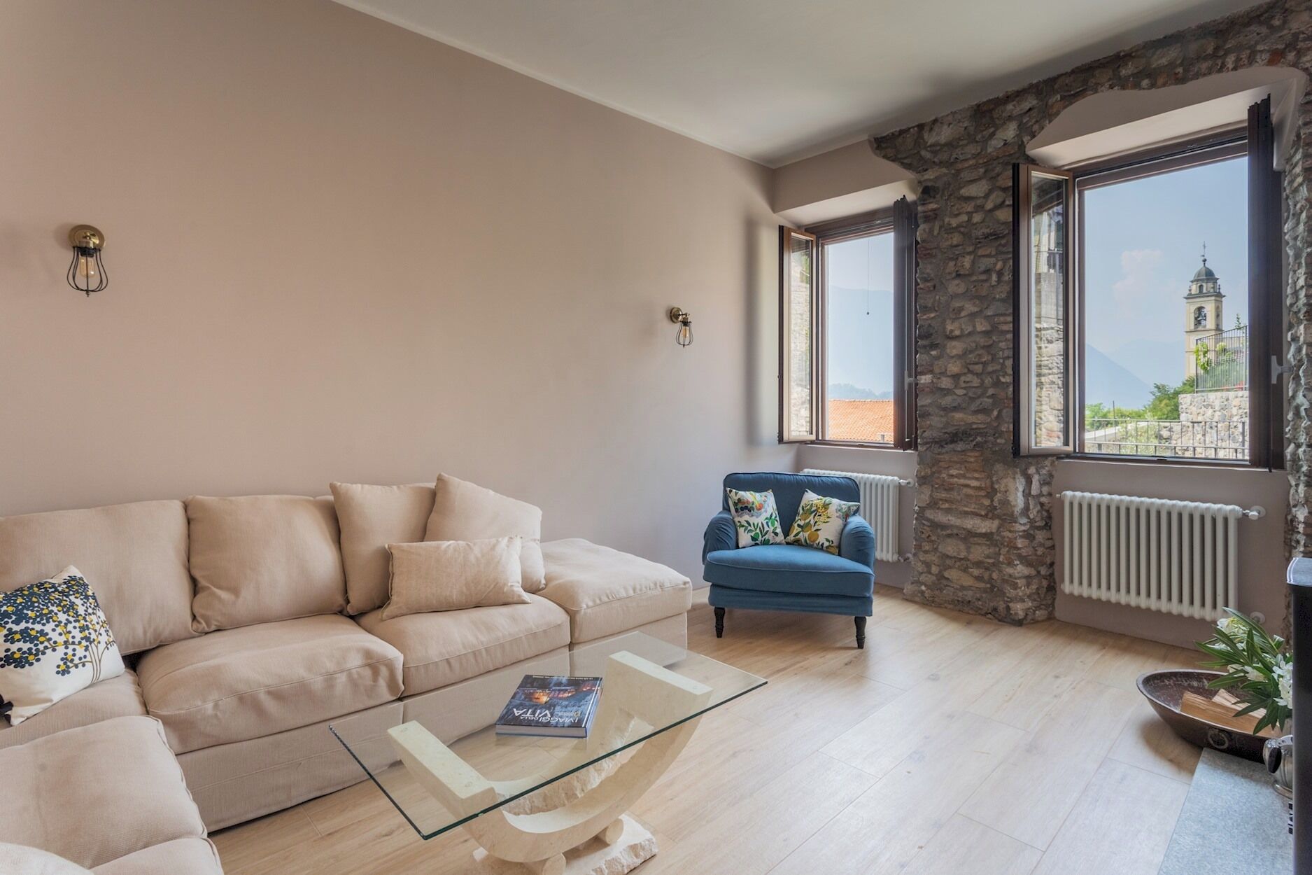 A living room with a beige sofa, a blue armchair, a glass coffee table, and a view of a tower through large windows.