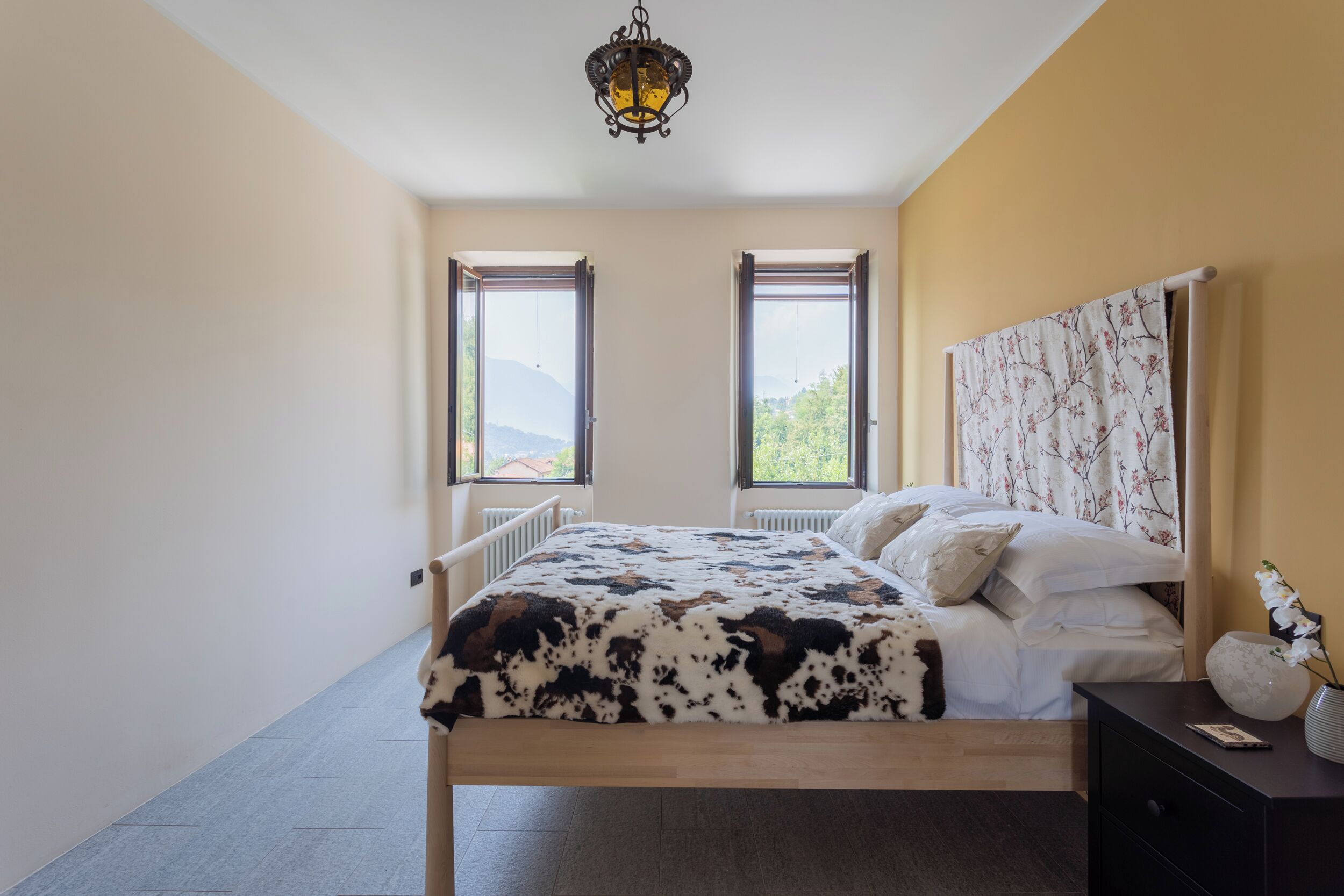 A bedroom with a bed, a nightstand, a lamp, and two windows.