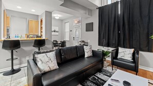 Ein modernes Wohnzimmer mit einer schwarzen Ledersofa, zwei schwarzen Barhocker, einer Essecke mit Tisch und Stühlen sowie einer Küchenzeile mit Arbeitsplatte und Geräten.