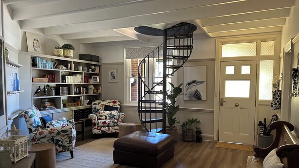 Een gezellige woonkamer met een houten vloer, een fauteuil met bloemenprint, een boekenkast, een spiltrap en een deur met een raam.