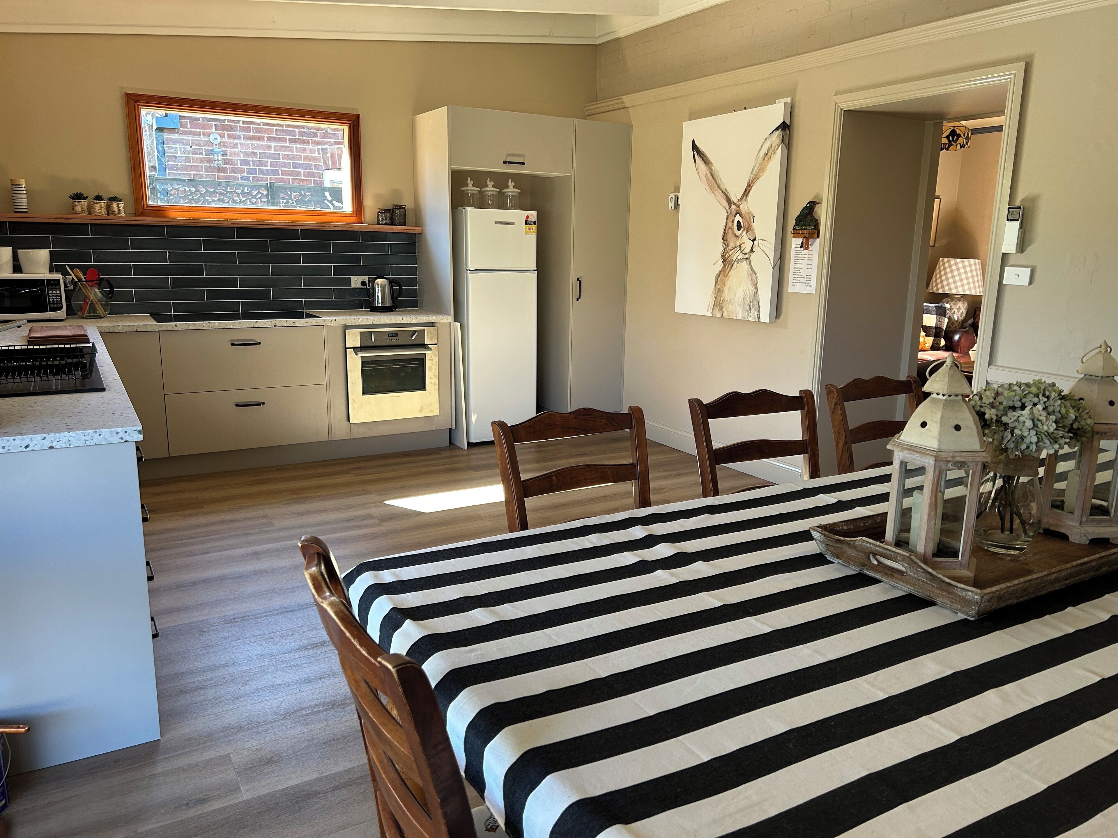 Una cocina moderna con horno empotrado, refrigerador y ventana con vista a un muro de ladrillo. Hay dos sillas de madera y una cama a rayas en la habitación.