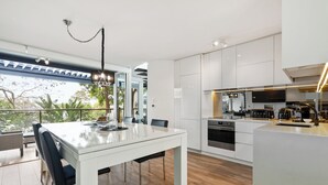 Una cocina moderna con armarios blancos, un comedor con mesa de vidrio y vistas a la vegetación exterior.