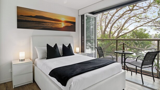 Un dormitorio moderno con una cama grande, una mesita de noche con lámpara, un balcón con mesa y sillas, y un cuadro de una puesta de sol.
