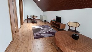 Una sala moderna con suelo de madera, un comedor con sillas, un televisor sobre un mueble de madera y un ventanal que deja entrar luz natural.
