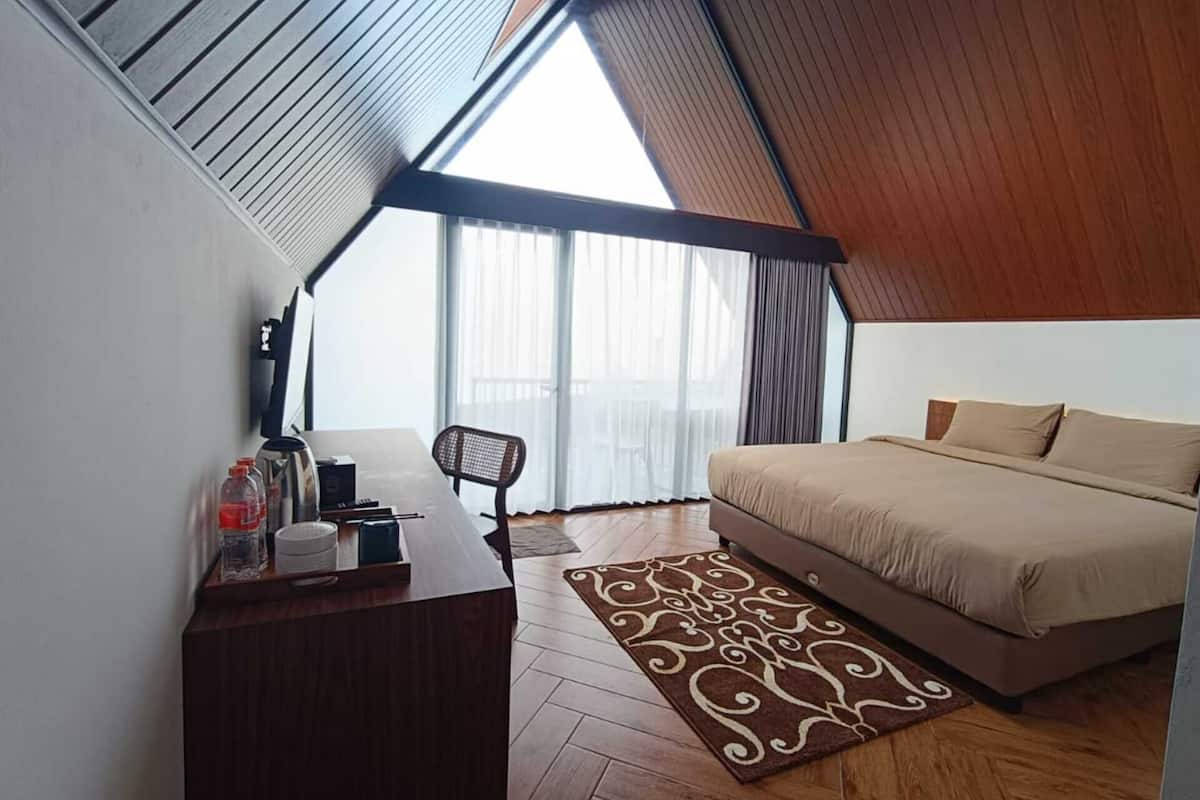 Une chambre avec un plafond en pente, un lit, un bureau avec une chaise et une grande fenêtre avec des rideaux.