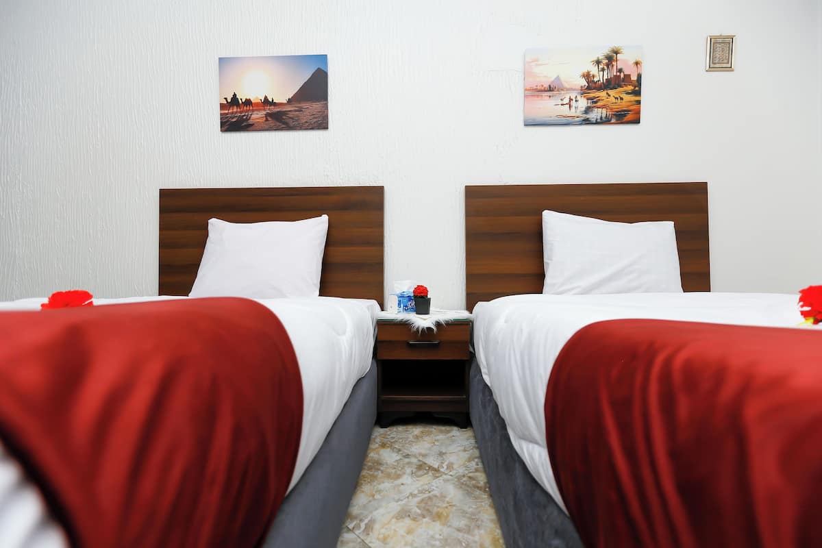 A hotel room with two beds, each with a wooden headboard, white bedding, and red throw blankets. There are two nightstands with small tables, a red flower, and a blue bottle. The walls are white, and there are two framed pictures above the beds.