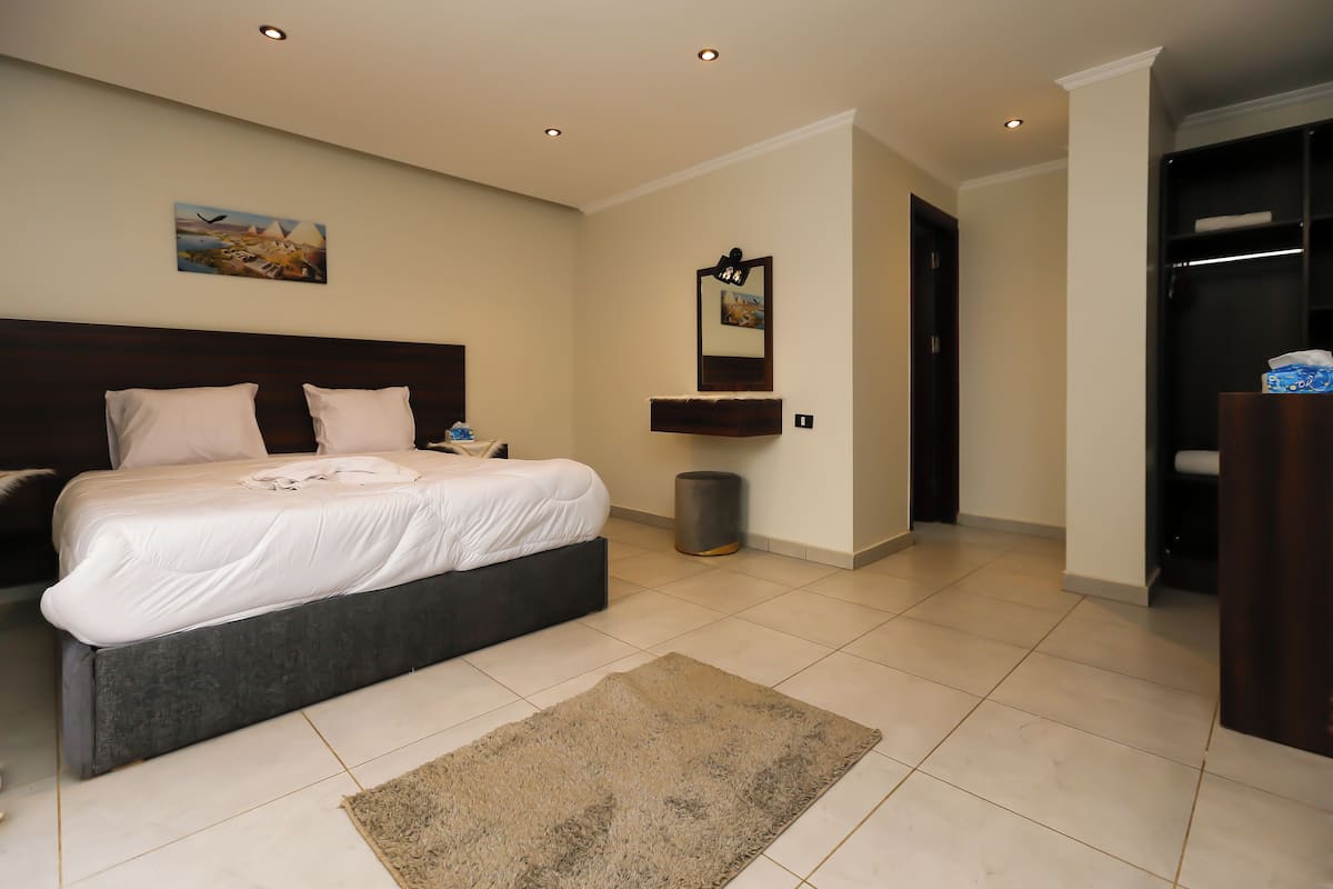 A modern bedroom with a large bed, a bedside table, a nightstand, a wall-mounted mirror, and a small trash can.