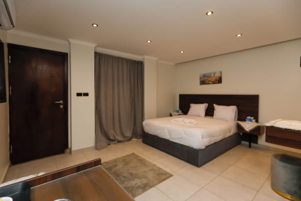 A hotel room with a bed, a desk, a chair, a trash bin, and a door.