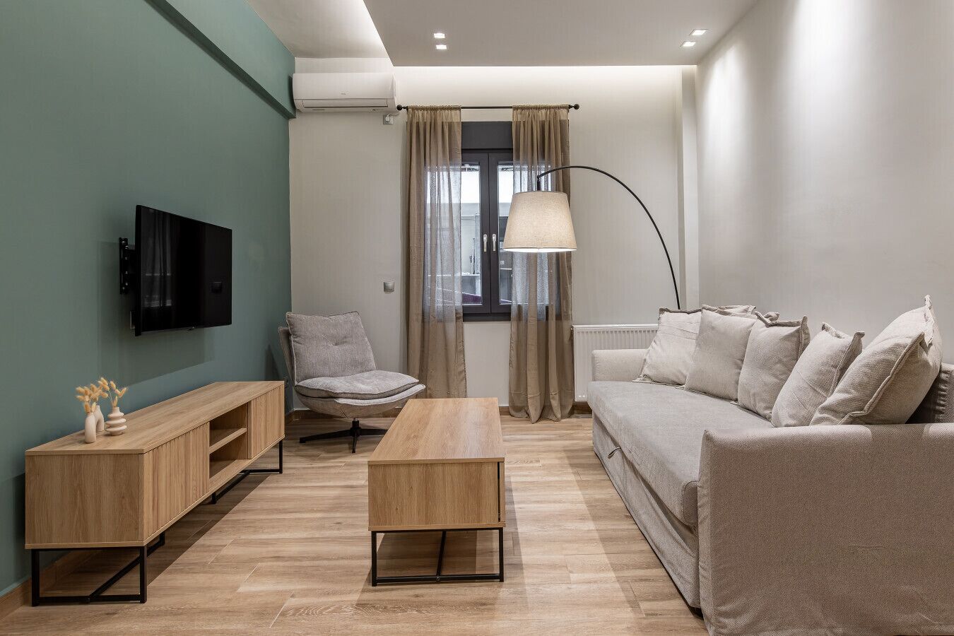 Ein modernes Wohnzimmer mit Flachbildfernseher, einem grauen Sessel, einem Holztisch, einem beigen Sofa und einer Stehlampe.