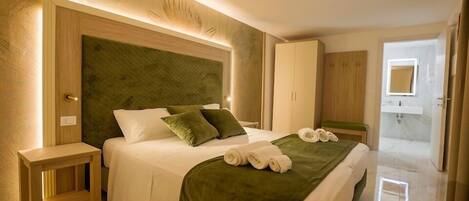 Una habitación de hotel moderna con una cama grande, cabecera de madera, mesita de noche y baño visible a través de una puerta abierta.