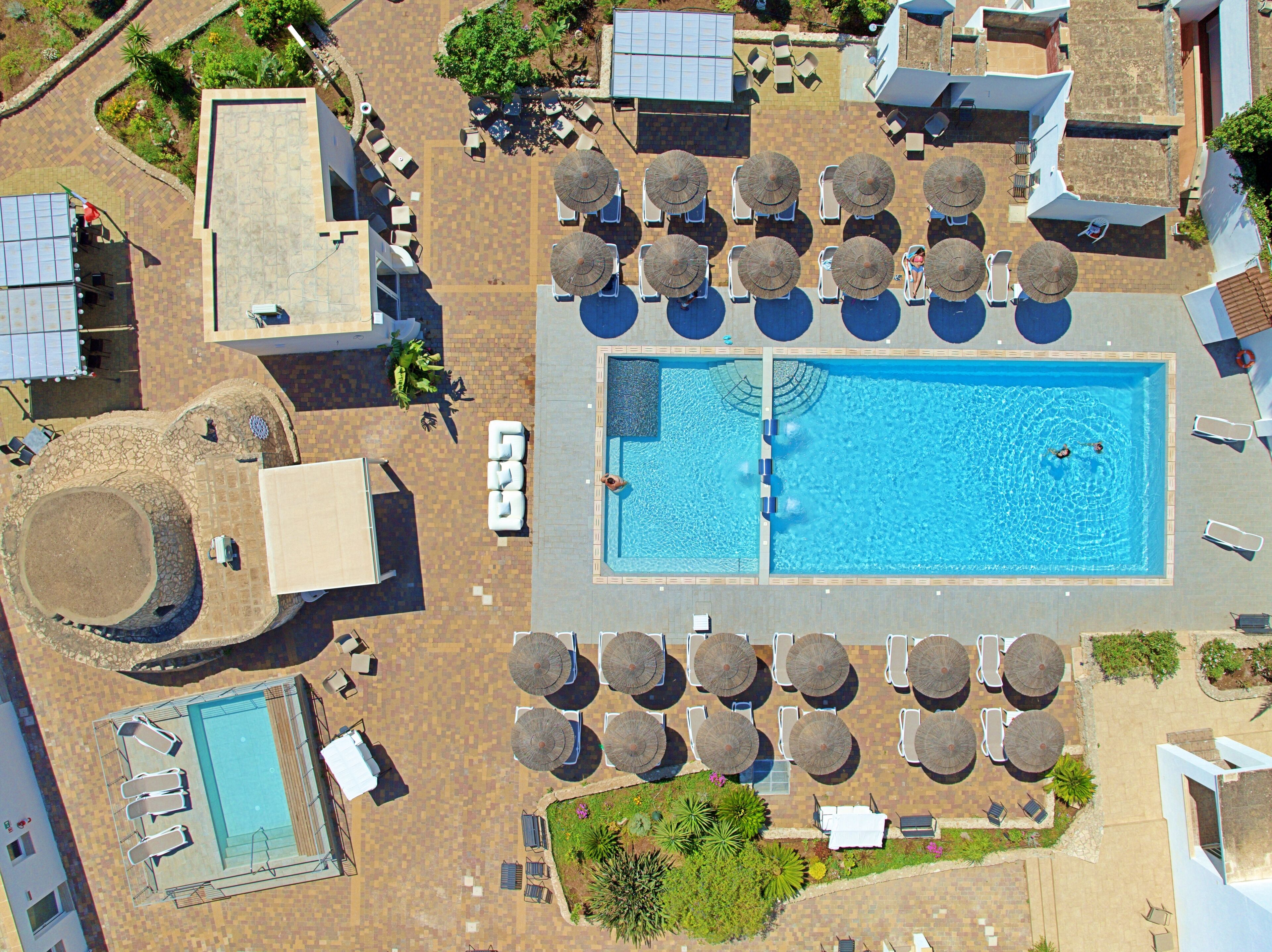 Vue aérienne d’un complexe hôtelier doté d’une piscine centrale, de nombreuses chaises longues et d’espaces ombragés.
