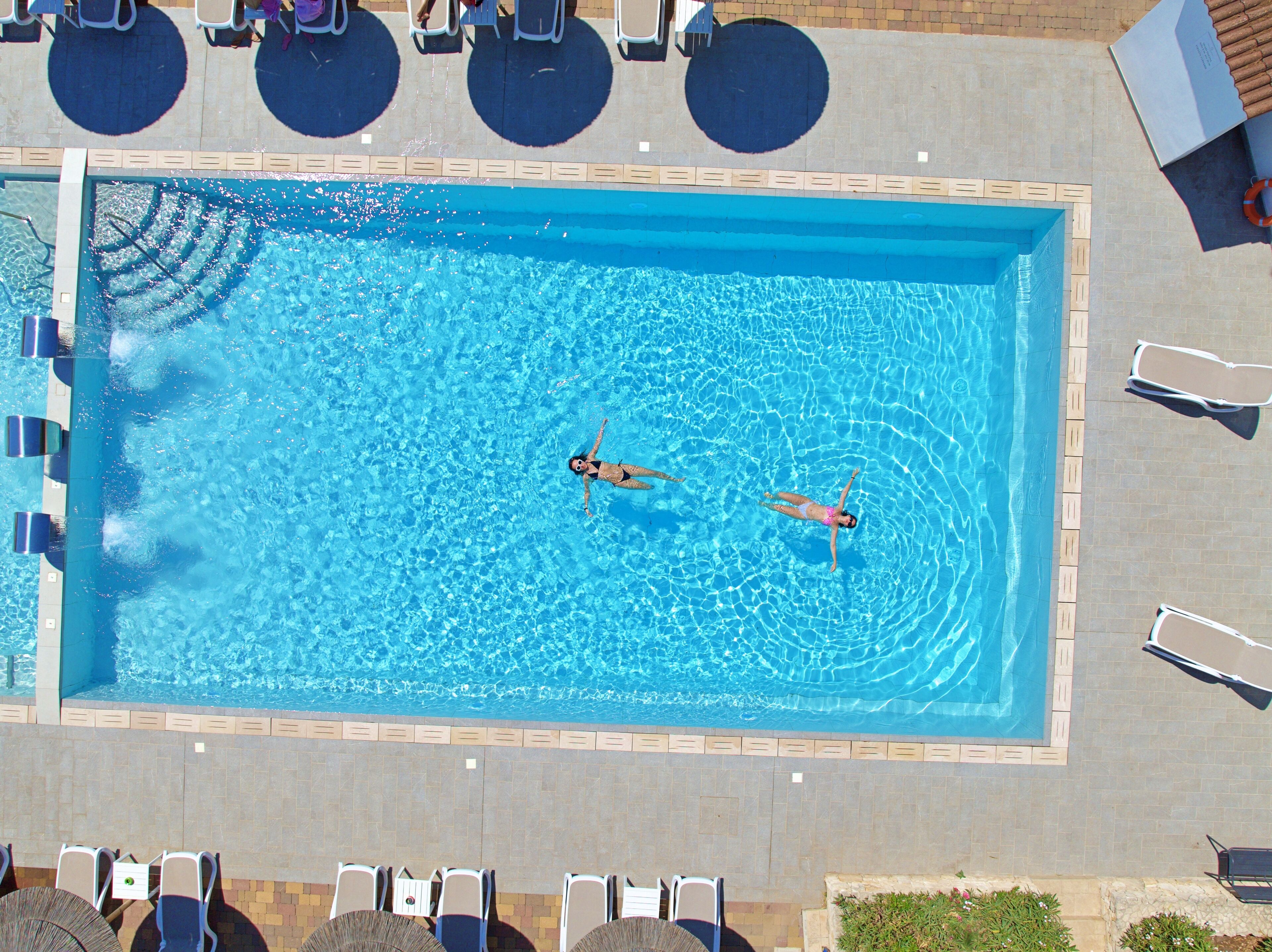 An aerial view of a rectangular swimming pool with two people swimming, surrounded by lounge chairs and umbrellas.