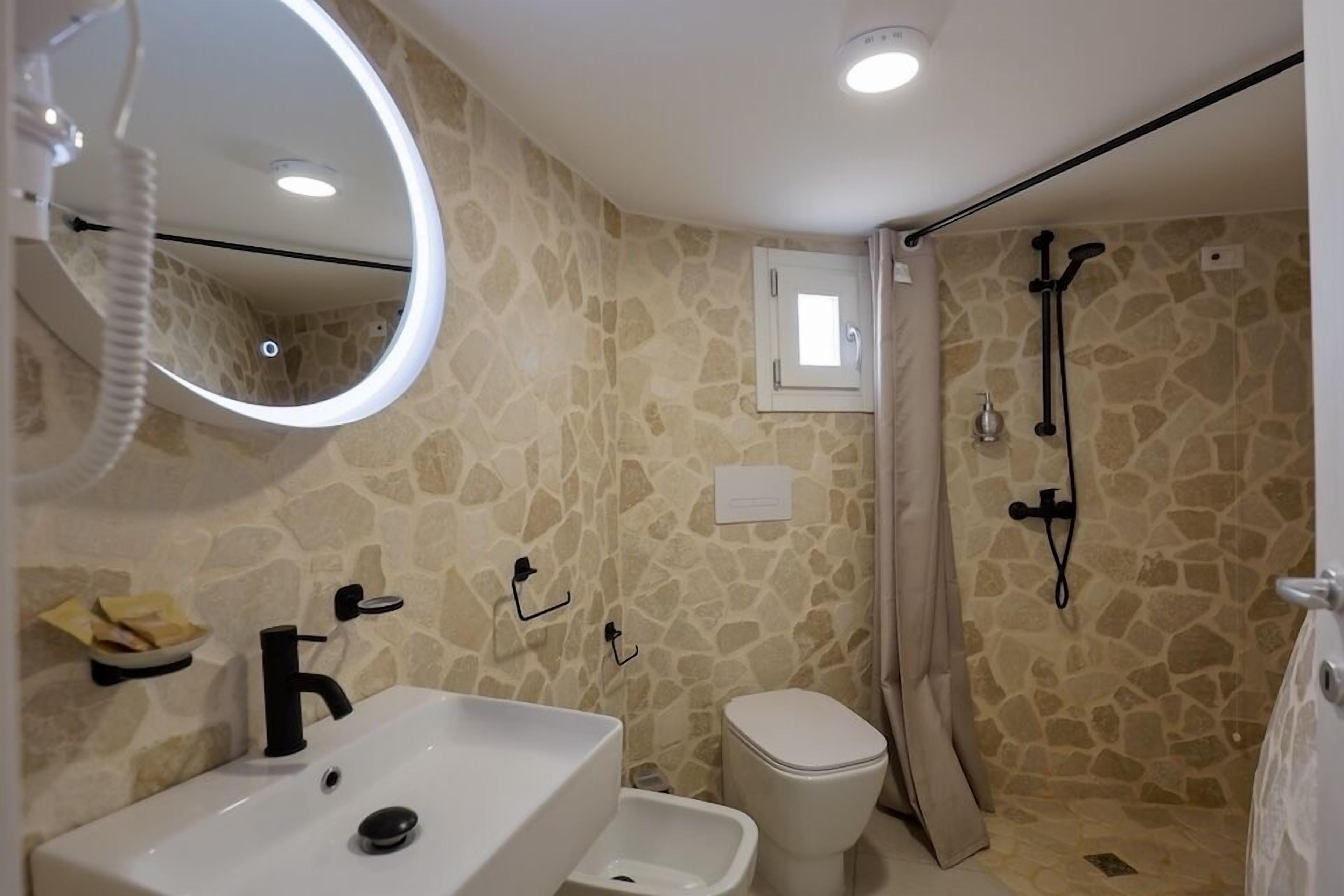 A bathroom with a stone wall, a white sink, a mirror, a toilet, and a shower area.