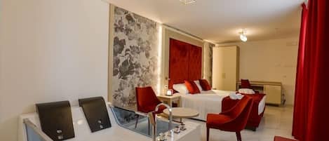 A hotel room with a large bed, a separate sitting area with a red chair, a small table, and a modern bathroom with a large bathtub and a mirror.