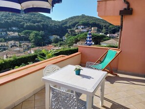 Ein Balkon mit einem weißen Tisch und Stühlen, einem blau-weißen Streifen-Sonnenschirm und einem grünen Klappstuhl.
