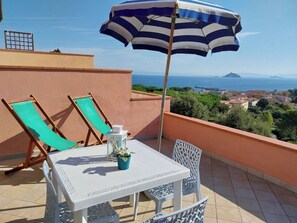 Una terraza con dos sillones verdes, una mesa blanca con una planta en maceta y un paraguas a rayas azul y blanco.