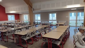 A spacious classroom with rows of tables and chairs, large windows, and a high ceiling with a perforated design.