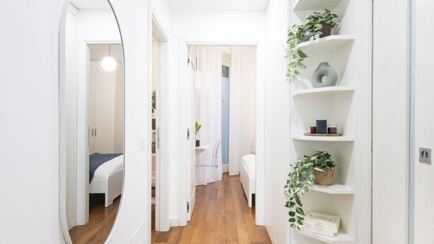 Een smalle gang met houten vloer, witte muren en een spiegelwand aan de linkerkant. Planten en decoratieve objecten staan op planken langs de rechtermuur.