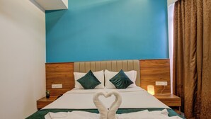 Habitación de hotel con cama, cabecero de madera, dos almohadas, mesita de noche con lámpara y un arreglo de toallas con forma de corazón sobre la cama.