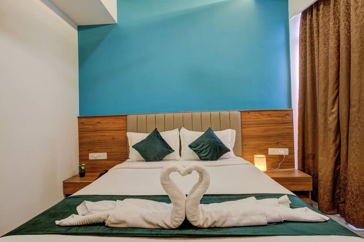 Une chambre d’hôtel avec un lit, une tête de lit en bois, deux oreillers, une table de chevet avec une lampe, et une disposition en forme de cœur réalisée avec des serviettes sur le lit.