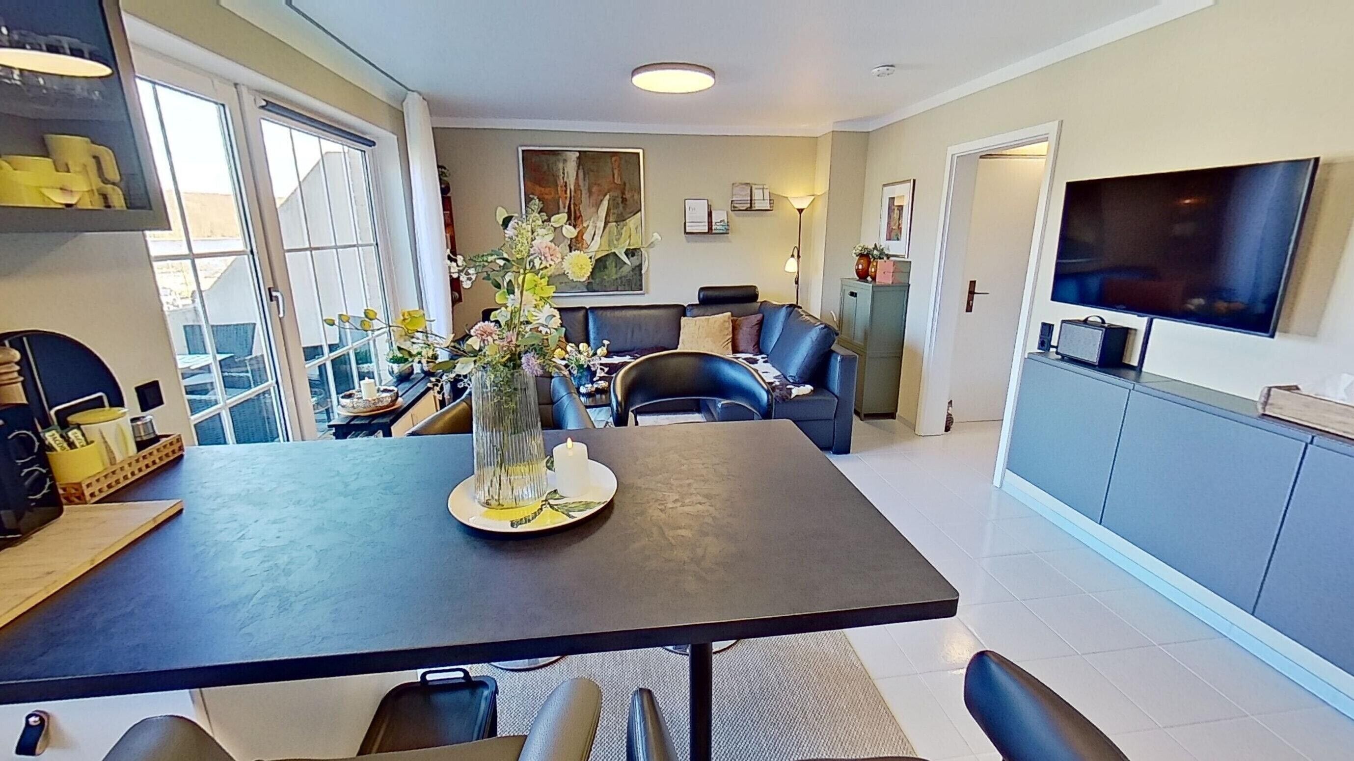 A modern kitchen with a dining table, chairs, a vase with flowers, a TV, and a cabinet.