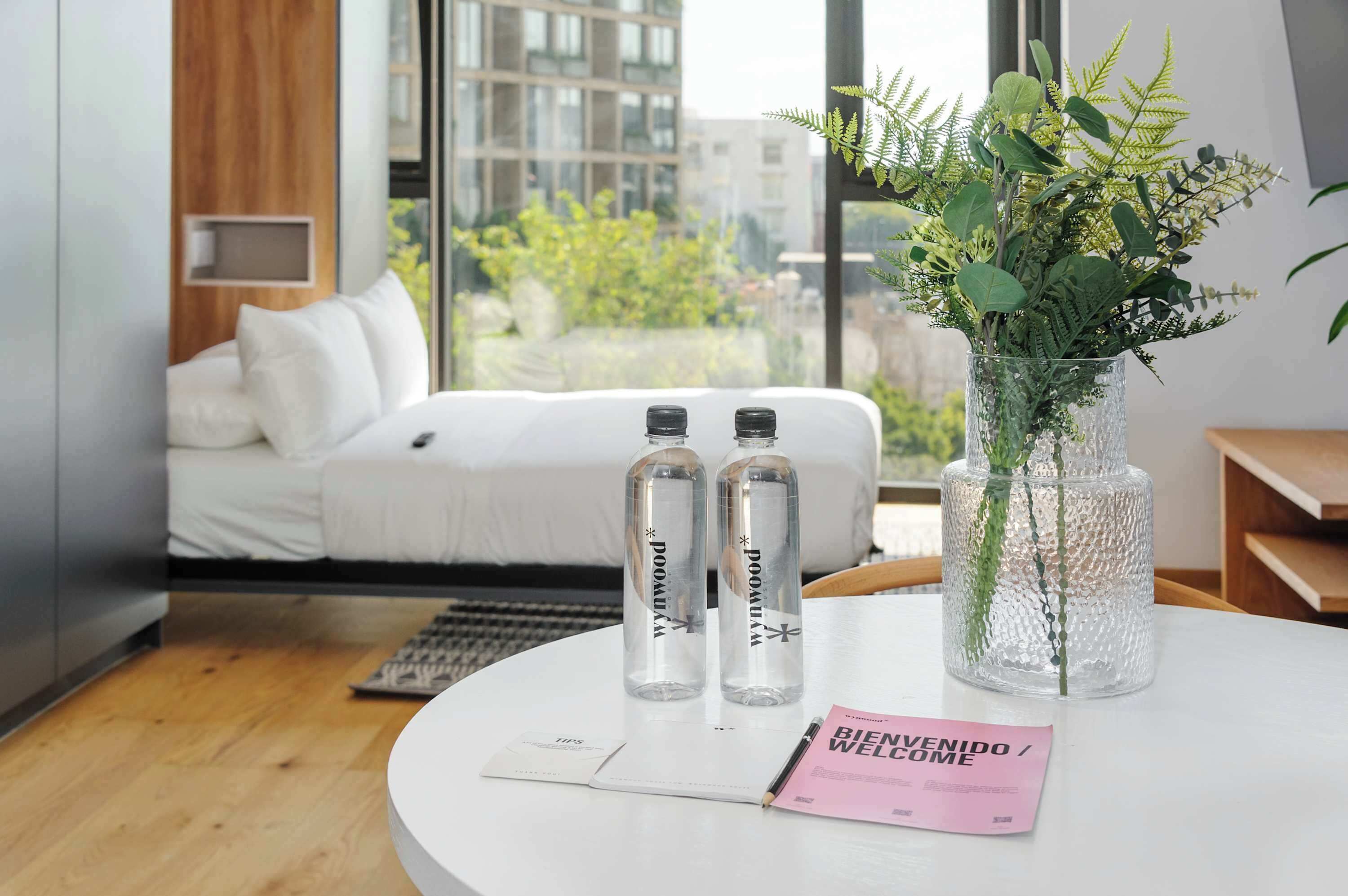 A modern hotel room with a bed, a round table with a vase of plants, and two bottles labeled "PODOMANYA".