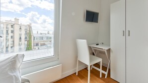 A small, bright room with a large window, a white desk, a chair, and a TV mounted on the wall. - Studio with panoramic view of the Eiffel Tower (Paris)