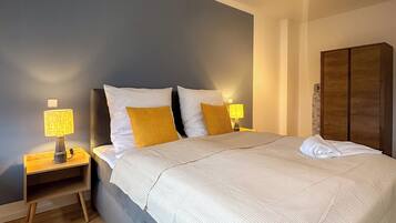Ein ordentlich gemachtes Bett mit weißer Tagesdecke und zwei Kissen, ein Nachttisch mit Lampe sowie ein Holzschrank in einem modernen Schlafzimmer.