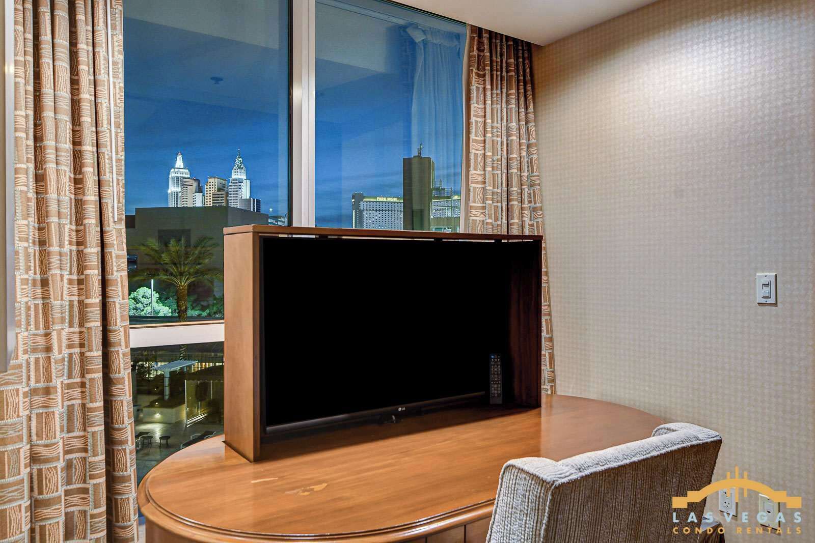 Habitación de hotel con un televisor de pantalla plana grande sobre un soporte de madera, una mesa de comedor con una silla y vistas a un paisaje urbano a través de amplios ventanales.