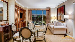Habitación de hotel con zona de comedor, sofá, televisor y ventana grande con vistas a la ciudad.