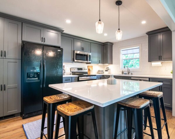 A modern kitchen with grey cabinets, a black refrigerator, a large island with stools, and pendant lighting. - Family Retreat in Birmingham – 7BR/4BA (Birmingham)