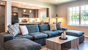 A modern living room with a sectional sofa, a coffee table, and a kitchen area with cabinets and appliances. - Family Retreat in Birmingham – 7BR/4BA (Birmingham)