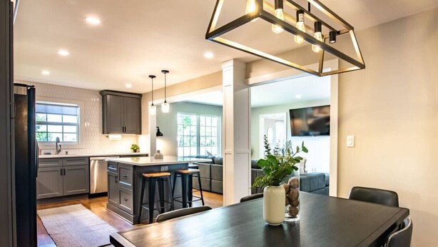 A modern kitchen with dark cabinetry, a central island with stools, and a dining area with a dark table and chairs. - Family Retreat in Birmingham – 7BR/4BA (Birmingham)