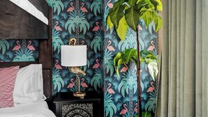 A bedroom with a bed, a nightstand with a lamp, and a large plant.