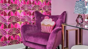 Ein violetter Samt-Sessel mit Kissen in Form eines Cupcakes, ein Glastisch mit Kristalllampe und eine Wand mit einem rosa und goldenen Fahnendruck.