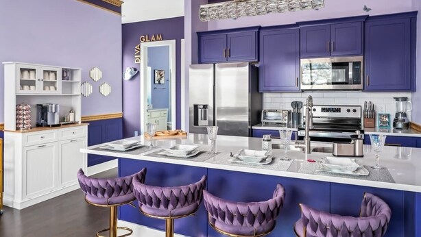 Eine moderne Küche mit violetten Schränken, einer zentralen Kochinsel mit Barhocker und einem Schild mit der Aufschrift 'DIVA GLAM'.