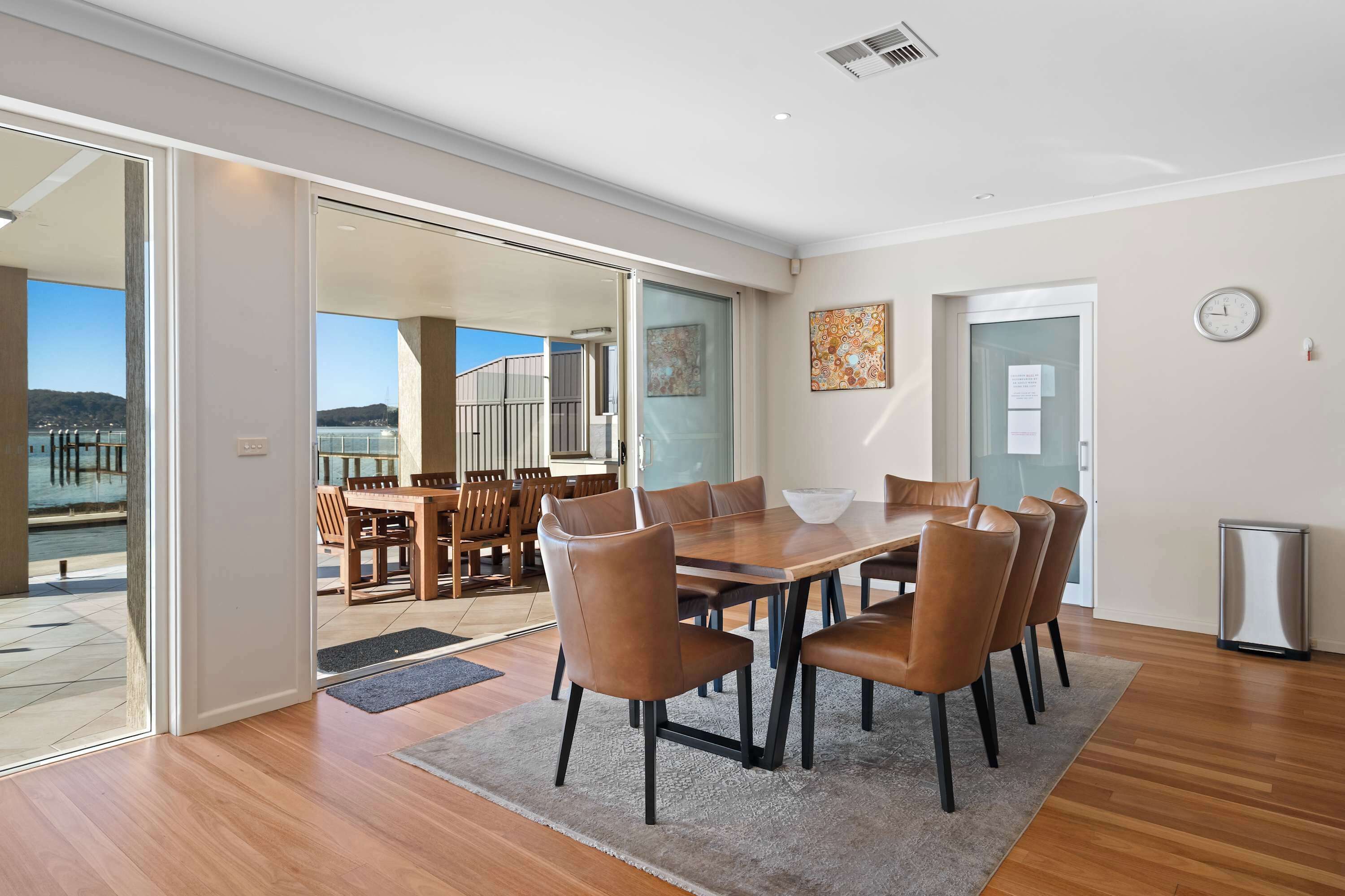 A spacious dining area with a large wooden table and leather chairs, a view of a balcony with a railing, and a clock on the wall.
